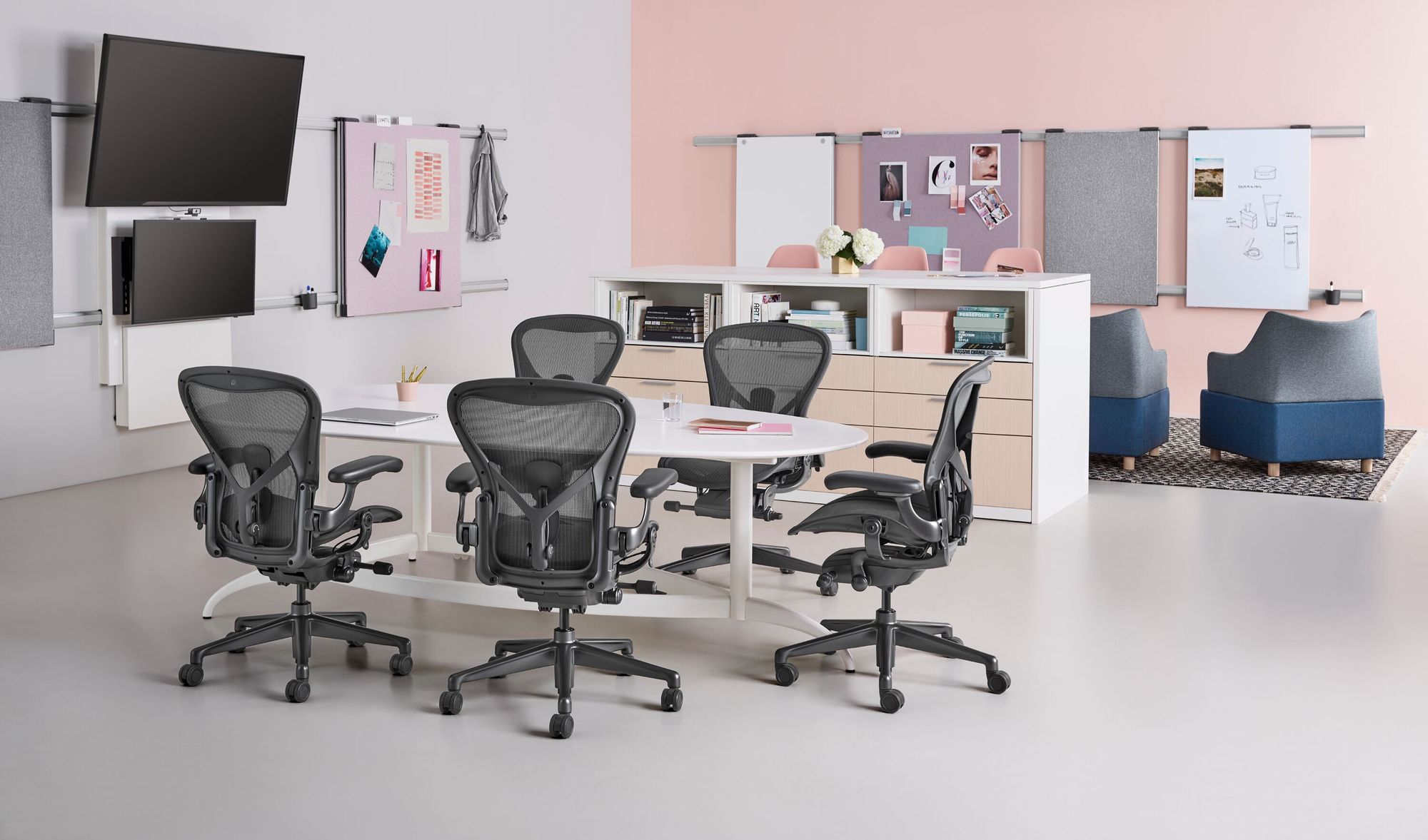Modernes Büro mit Aeron Stühlen von Herman Miller, die um einen runden weißen Tisch angeordnet sind. Die organisierte Arbeitsfläche ist in sanften Rosa- und Grautönen gehalten, mit Akzenten durch blaue Loungesessel und strukturierte Trennwände. Stilvolle Organisationselemente und eine minimalistische Dekoration ergänzen die ästhetische und ergonomische Raumgestaltung